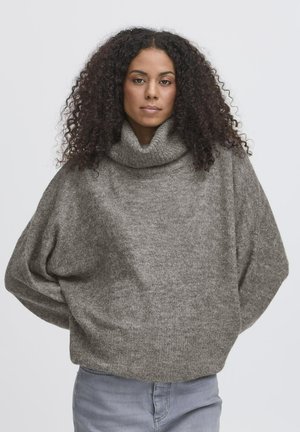 Femme aux cheveux bouclés portant un pull à col roulé gris ample et un jean bleu clair, face à l'avant, les mains dans le dos.