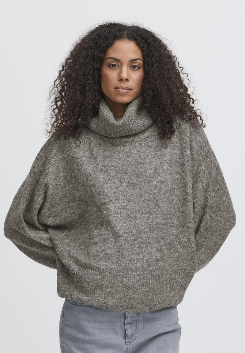 Femme aux cheveux bouclés portant un pull à col roulé gris ample et un jean bleu clair, face à l'avant, les mains dans le dos.