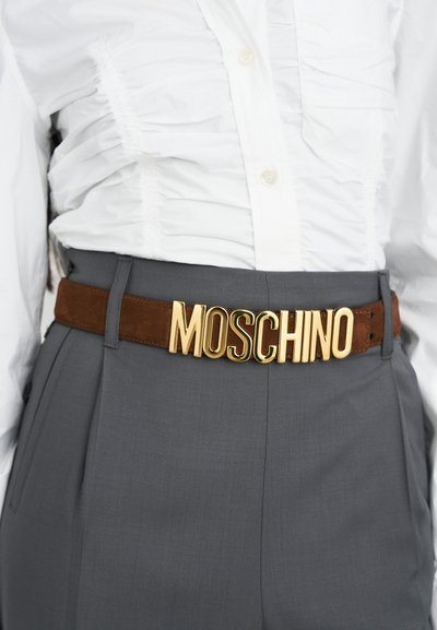 Brown suede belt with large gold lettering spelling "MOSCHINO," worn over a white gathered shirt and gray trousers.