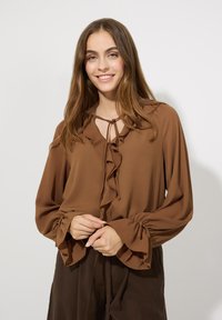 Braune Bluse mit Rüsche am Ausschnitt und an den Ärmeln, aus einem weichen Stoff gefertigt. Verfügt über Kordeln am Hals und geraffte Bündchen.