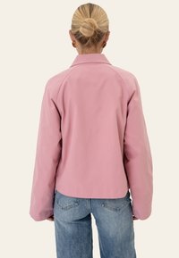 Femme aux cheveux blonds en chignon, vêtue d'une veste rose et d'un jean bleu, vue de dos sur un fond uni.