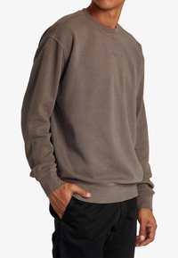 Sweat-shirt gris avec texture minimale, poignets et ourlet côtelés ; présente un logo discret au centre. Associé à un pantalon noir.