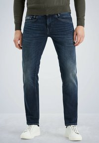 Jean en denim bleu foncé avec une finition légèrement délavée, coupe droite, design à cinq poches et une étiquette sur la poche arrière. Associé à des baskets blanches.