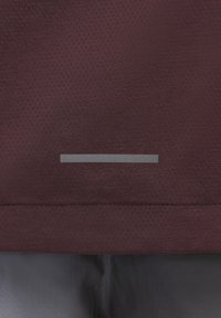 Nahaufnahme von dunkelrotem, durchlöchertem Stoff mit einem horizontalen, grauen reflektierenden Streifen über einer dunkelgrauen Hose.