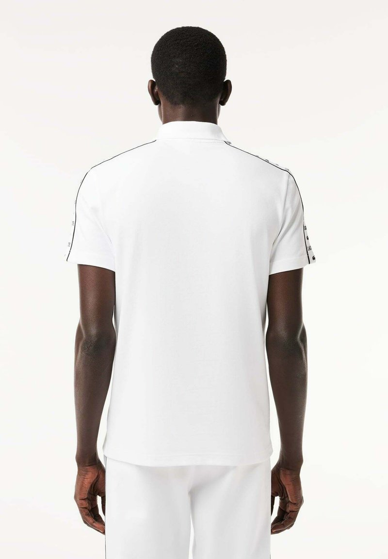 Chemise polo blanche à manches courtes, avec des détails noirs le long des épaules, texture lisse et col classique.