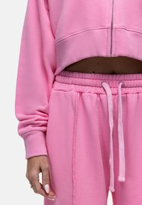 Felpa rosa corta con maniche lunghe e zip, abbinata a pantaloni della tuta rosa coordinati con cintura elastica e lacci. 