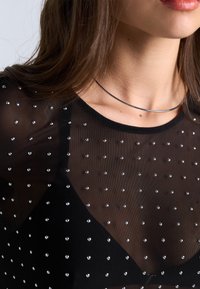Gjennomsiktig svart topp med spredte sølvstrass, rund halslinning og en glatt metallikk choker. Stoffet er lett og transparent.