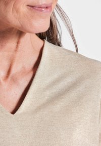 Beige v-hals top van een glanzende, gestructureerde stof. Close-up benadrukt de halslijn en het zachte, gladde materiaal. Licht en subtiel glanseffect.