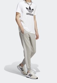 Valkoinen t-paita, jossa on musta Adidas-logo, vaaleanharmaat collegehousut valkoisilla sivuraitoilla ja beigewi sneakersit mustilla yksityiskohdilla.