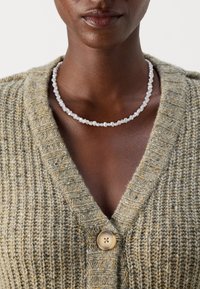 Pärlhalsband med oregelbundna former och storlekar, vit färg, bärs över en texturerad, olivgrön cardigan med V-hals och knappdetalj.