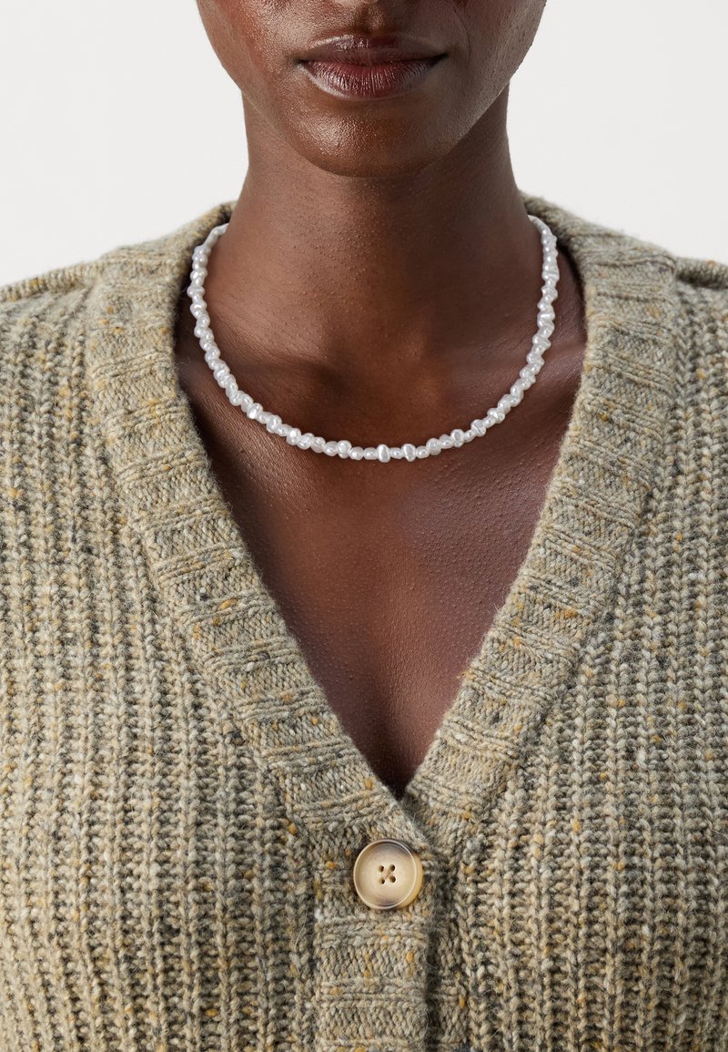 Pärlhalsband med oregelbundna former och storlekar, vit färg, bärs över en texturerad, olivgrön cardigan med V-hals och knappdetalj.