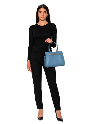 Frau in einem schwarzen Langarmoberteil und einer Hose, die eine mittelblaue Handtasche hält und schwarze High Heels trägt, vor einem weißen Hintergrund.