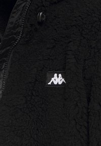 Giacca in pile nera con superficie texture, chiusura con zip e una toppa bianca con logo sul lato sinistro.