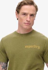 Oliven grøn bomulds t-shirt med rund halsudskæring og korte ærmer, med "superdry" logo i kontrasterende gult på brystet.
