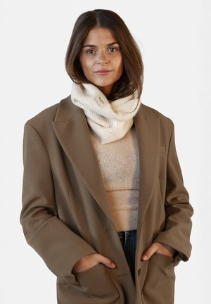 Sciarpa infinita in maglia beige indossata con un blazer oversize marrone chiaro, caratterizzata da un motivo testurizzato e un'etichetta cucita di lato.