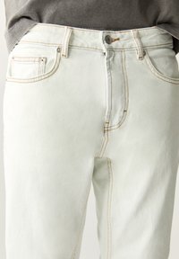 Lichtgroene spijkerbroek met een aangesloten snit, voorzien van beige stiksels, voorzakken en een metalen knoopsluiting bij de taille.