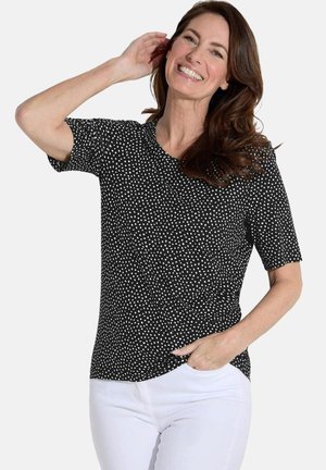T-shirt noir à manches courtes avec des pois blancs, encolure ronde, tissu lisse et coupe décontractée. Le modèle présente un style décontracté avec un pantalon blanc.