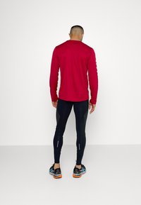 T-shirt de sport rouge à manches longues, legging noir ajusté et chaussures de course noires avec des accents orange, vu de dos. Texture de tissu lisse.