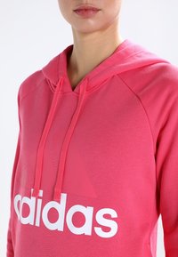 Rosa hoodie med huva och vit "adidas"-logotyp samt triangeldesign. Tillverkad av mjukt material, med snoddar och raglanärmar.