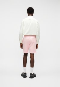 Άνδρας που φοράει ανοιχτό ροζ σορτς, λευκό μακρυμάνικο πουκάμισο μέσα από το παντελόνι, λευκές κάλτσες και μαύρα αθλητικά παπούτσια, όρθιος με την πλάτη στην κάμερα.