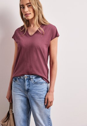 Jonge vrouw die een stofroze T-shirt met V-hals en lichtblauwe jeans draagt, een beige tas vasthoudend tegen een effen lichte achtergrond.