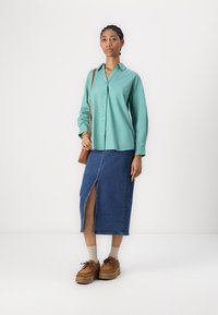 Camicia turchese a bottoni, gonna di jeans con spacco laterale, scarpe brown con zeppa e borsa a tracolla beige. Tessuto morbido, vestibilità casual.