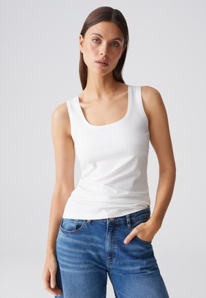 Woman with long brown hair wearing a white tank top and blue jeans standing with one hand in pocket against a plain background.