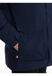 Tmavě modrá nepromokavá bunda s manžetou na suchý zip, bočními kapsami a štítkem s logem. Disponuje hladkou texturou a knoflíkovými uzávěry.