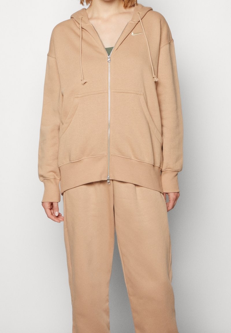 En person som bär en beige Nike zip-up hoodie och matchande beige sweatpants, som står mot en enfärgad vit bakgrund.