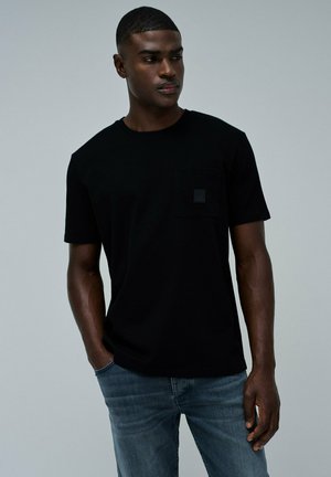 Camiseta negra de manga corta con cuello redondo y un pequeño bolsillo cuadrado en el pecho. Fabricada con una tela suave y lisa, presenta un corte relajado.