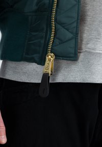 Prešívaná zelená bomber bunda so zlatým zipsom, s šedým rebrovaným pásom a čiernymi nohavicami. Viditeľné textúry a stehy.