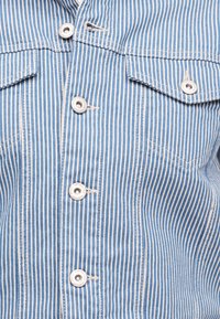 Ύφασμα με κάθετες ρίγες σε μπλε και άσπρο, με ασημένια κουμπώματα και δύο εμπρόσθιες τσέπες με καπάκι σε ένδυμα.