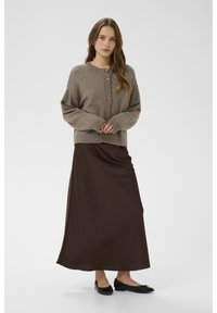 Ruda megzta kardiganas su sagomis, derinamas su ilga ruda satino suknele ir juodais bateliais be kulno. Modelis stovi sukryžiavęs rankas.