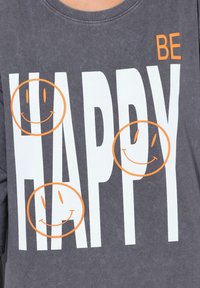 Grijze T-shirt met grote witte tekst "WEES VREDELIJK" en drie oranje glimlachende gezichten. Ronde halslijn en zachte, gestructureerde stof.