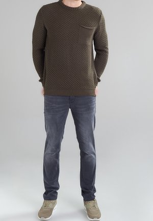 Olivgrüner Strickpullover mit strukturiertem Muster, Rundhalsausschnitt und Tasche; kombiniert mit grauen Jeans und beigen Sportschuhen. Neutraler Hintergrund.