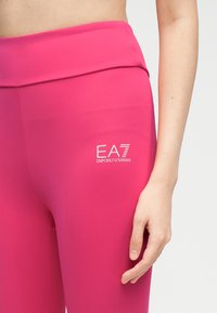 Γυναίκα που φοράει έντονο ροζ κολάν με το λογότυπο Emporio Armani EA7 κοντά στο ισχίο, με το χέρι χαλαρά στο πλάι.