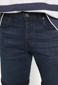 Mörkblå denimjeans har en slät yta, femficksdesign och metallknappstängning. Sömmen är uppvikt för en croppad effekt.