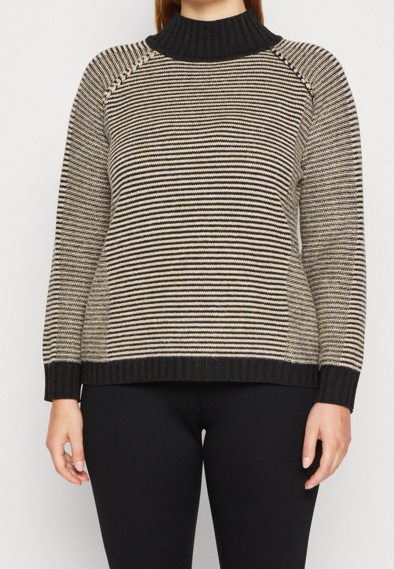 Femme portant un pull en tricot beige et noir à rayures avec un col roulé côtelé noir, des poignets et un ourlet, associé à un pantalon noir.