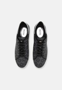 Zapatillas negras con una tela texturizada que presenta un patrón repetido del logo de MK, cordones negros y una suela de goma blanca en contraste.