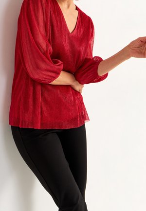 Kobieta ubrana w czerwoną teksturowaną bluzkę z dekoltem w serek i bufkami na rękawach oraz czarne spodnie, pozująca na białym tle.