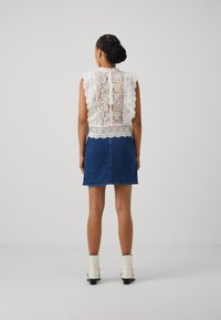 Weißes Spitzenoberteil mit wellenförmigen Kanten, darüber ein eng anliegender blauer Jeansrock. Das Modell trägt weiße Stiefeletten mit klobigen Absätzen.