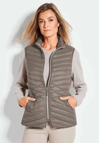 Gepatchte graue Weste mit Stehkragen, Vorderreißverschluss und seitlichen Taschen, über einem hellbeigen Pullover getragen. Glatte Stoffoberfläche.