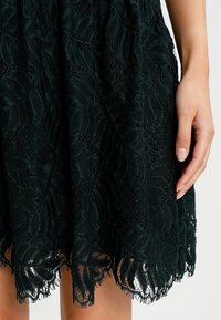 Dunkelgrünes Spitzenkleid mit floralem Muster, das einen strukturierten, fransigen Saum aufweist. Der Stoff ist halbtransparent und schichtet sich für zusätzlichen Tiefeneffekt.
