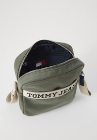 Borsa a tracolla verde in tela con chiusura zip. Presenta un'etichetta in tessuto "TOMMY JEANS" e un interno navy con una piccola tasca zip.