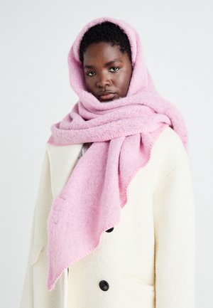 Femme portant une écharpe rose pâle enroulée autour de la tête et un manteau blanc avec des boutons noirs, sur un fond clair uni.