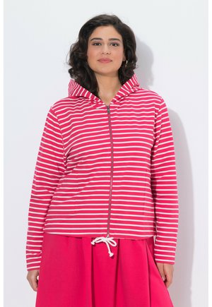 Femme portant un sweat à capuche zippé rouge et blanc à rayures avec une capuche, et une jupe rouge unie avec un cordon de serrage blanc.