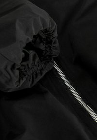 Gros plan sur la manche d'une veste noire avec un poignet élastique et une fermeture éclair argentée visible sur un fond en tissu noir.