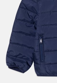 Giacca puffer blu navy con design trapuntato, dotata di tasca con zip sulla manica e polsini elastici per una vestibilità aderente. Tessuto dalla texture liscia.