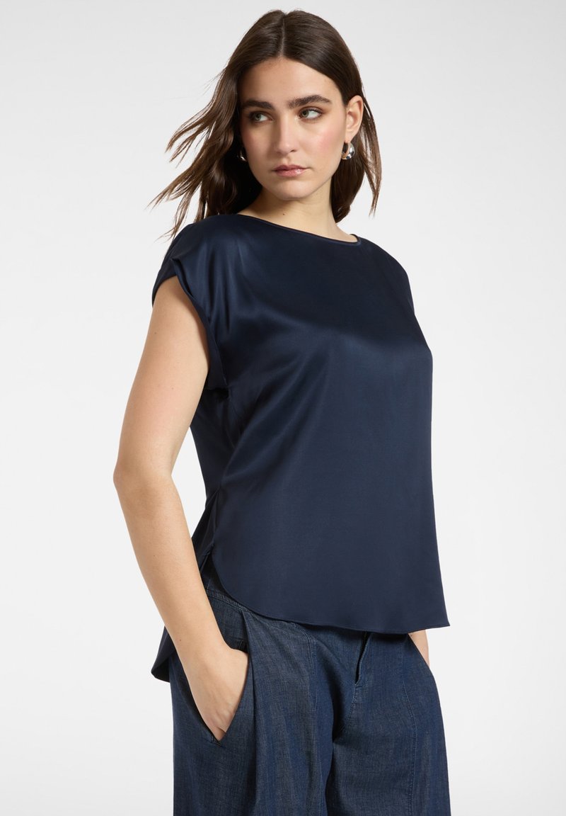 Mujer con cabello oscuro suelto, que lleva pendientes de aro plateados y una blusa de satén azul marino de manga corta, con pantalones azul oscuro, con las manos en los bolsillos.