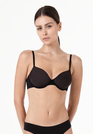 Donna con capelli scuri raccolti in una coda bassa, che indossa un reggiseno nero con ferretto e mutandine nere abbinate, su uno sfondo bianco.